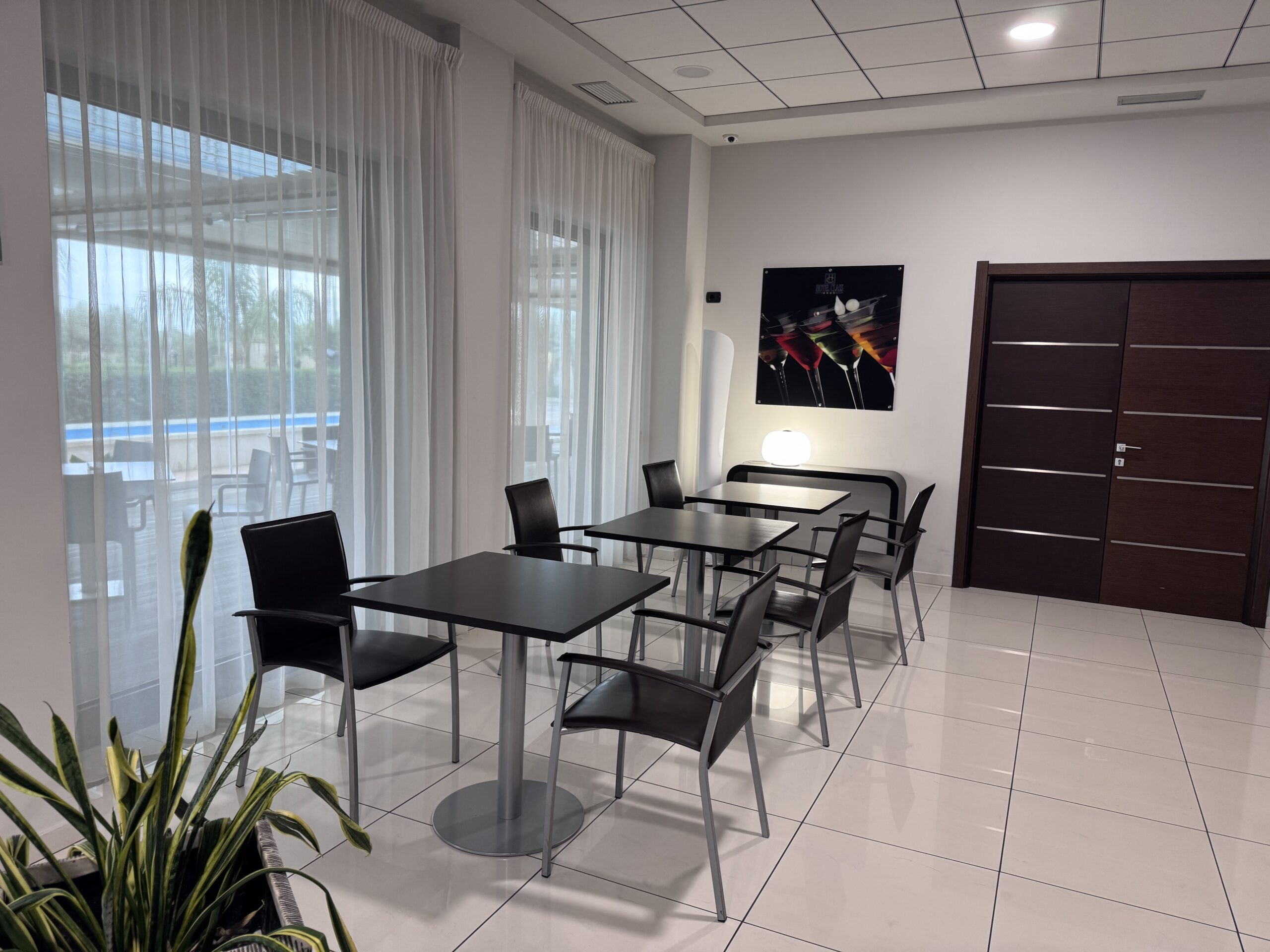 Bright interior room with modern minimalist tables and chairs, large windows with white curtains that let in natural light, ornamental plant in the foreground, colorful abstract painting on the wall, and light-colored tile floor.
