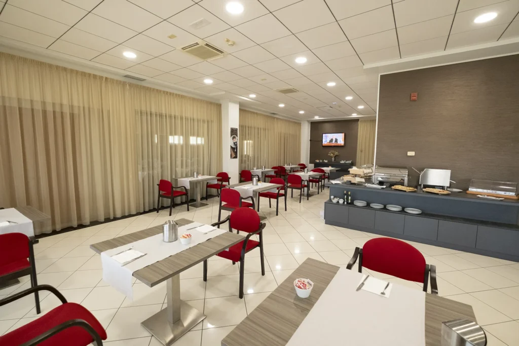 Hotel breakfast room with wooden tables set with white tablecloths, red upholstered chairs, and light-colored tile flooring; in the background, a buffet with food and beverages, walls with beige curtains, and a wall-mounted television in a bright setting.