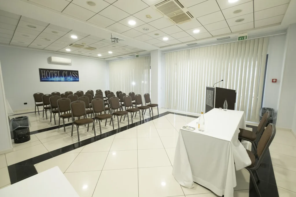 Hotel meeting room with speaker's table covered with a white tablecloth, upholstered chairs and microphone, rows of chairs arranged for the audience, light-colored walls with vertical panels and “Hotel Class” logo; bright room with polished tile floor and ceiling projector.