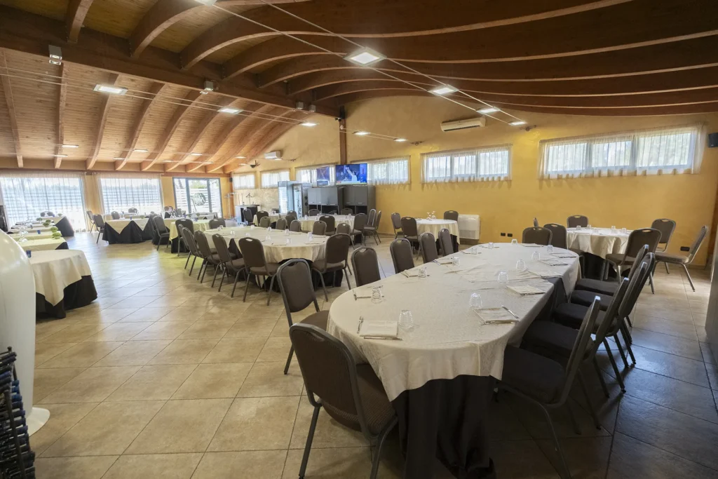 Spacious restaurant with curved wooden beams, round and rectangular tables set with white tablecloths and glasses, neatly arranged upholstered chairs, large windows letting in natural light, wall-mounted televisions, and a welcoming atmosphere ideal for breakfast, lunch, and dinner at the hotel.