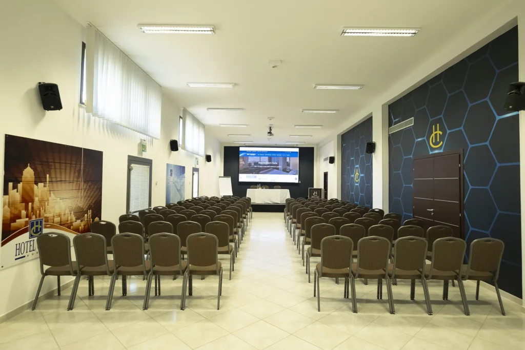 Spacious conference room with rows of chairs neatly arranged facing a large projection screen, modern walls with blue geometric decorations and the hotel logo, uniform ceiling lighting, light-colored tile flooring, and a professional atmosphere ideal for meetings and corporate events.